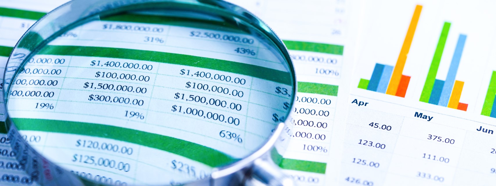 A magnifying glass placed over a printout of a spreadsheet showing dollar amounts and a chart of monthly figures.