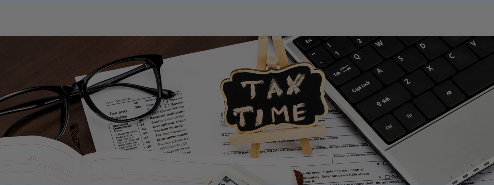 Closeup of tax forms, eyeglasses, laptop, and TAX TIME written on tiny chalkboard