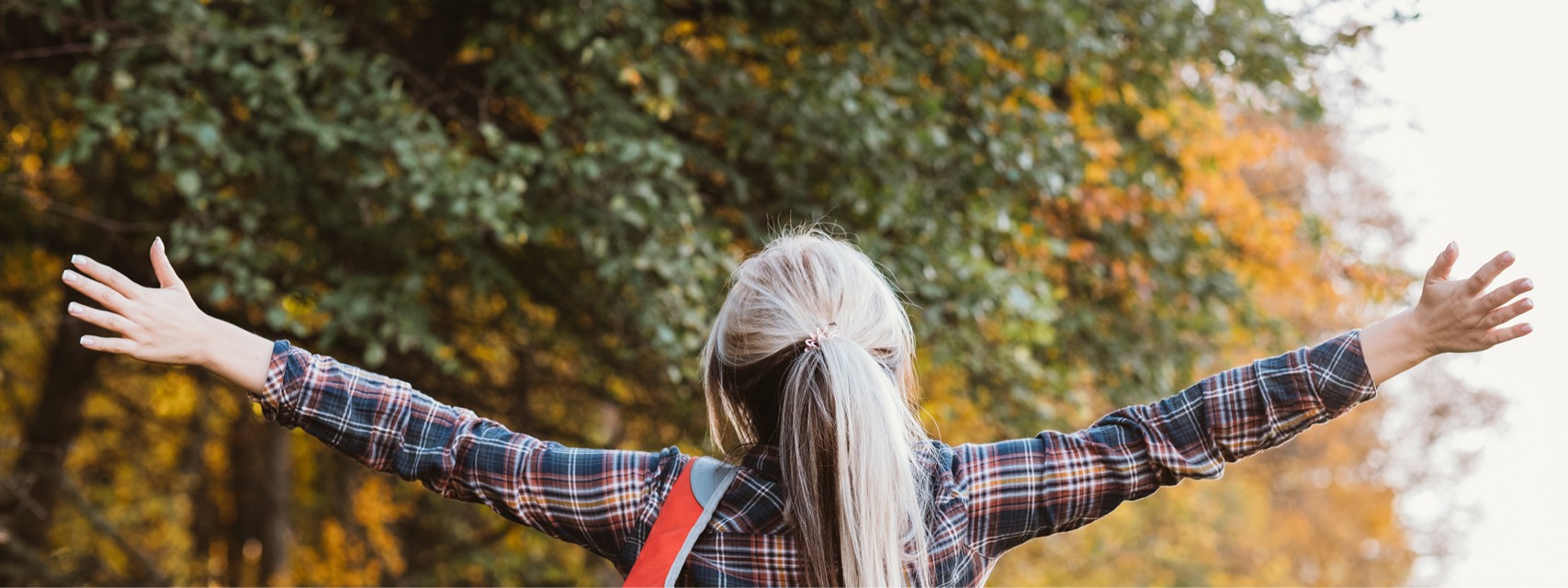 Lady outdoors with arms stretched wide, surrounded by autumn-colored trees.